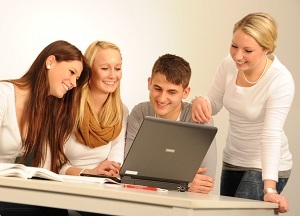 Schmuckgrafik (Beispielbild) "Drei sitzende Personen vor einem Laptop und einer daneben stehenden Frau"